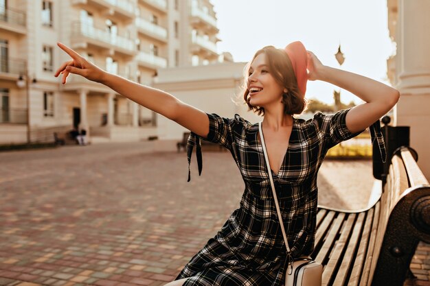 Chica romántica delgada sentada en un banco con una sonrisa. Tiro al aire libre del dedo acusador modelo femenino francés extático en algo.