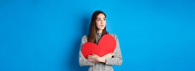 Chica romántica del día de san valentín mirando soñadora en la esquina superior izquierda y sonriendo sosteniendo un gran corazón rojo c