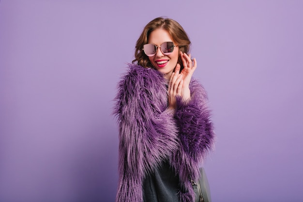 Chica romántica en elegante traje morado posando con sonrisa tímida en estudio