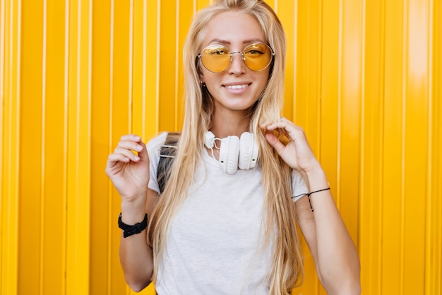 Chica romántica en gafas de sol amarillas posando con sonrisa interesada sobre fondo brillante. chica bronceada con largo cabello rubio riendo a la cámara.