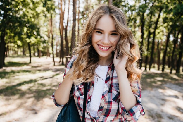 Chica romántica hablando por teléfono mientras posa en el bosque. Foto al aire libre de elegante mujer rubia con alegre sonrisa de pie en la naturaleza.