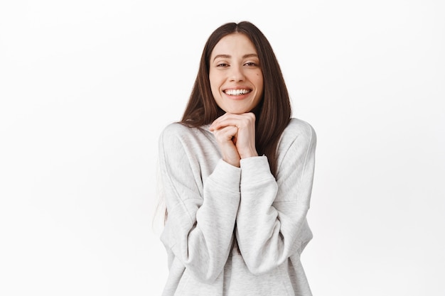 Chica romántica y linda que mira con admiración y simpatía, admira algo, observa una hermosa escena encantadora, apoya la cabeza en las manos y sonríe, de pie sobre una pared blanca