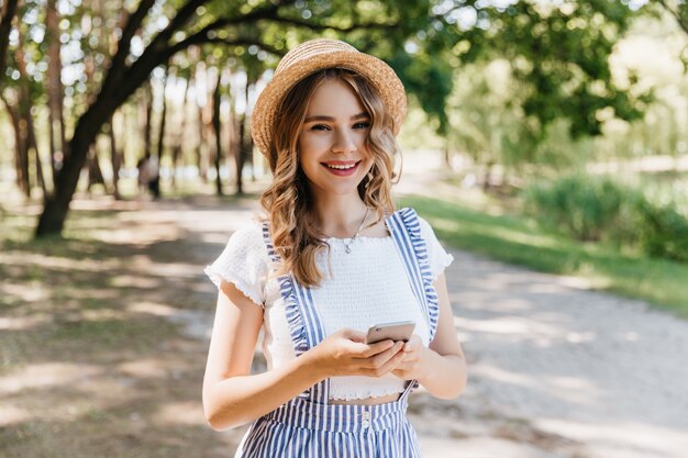 Chica romántica lleva sombrero y camiseta blanca sonriendo a la naturaleza. Adorable mujer rubia disfrutando de un paseo en el parque.