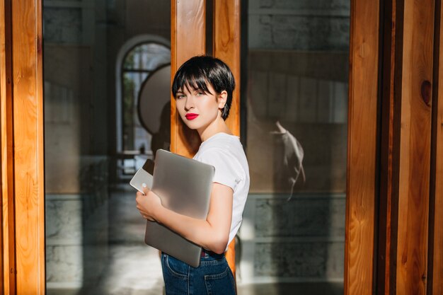 Chica romántica con peinado corto de pie cerca de la puerta y sosteniendo una computadora portátil y un teléfono inteligente