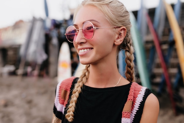 Chica romántica con peinado de moda posando en gafas de sol rosas. Tiro al aire libre de hermosa dama rubia con trenzas disfrutando del buen tiempo.