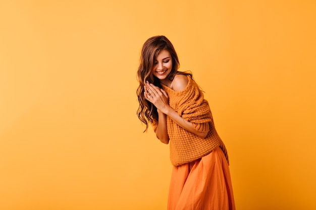 Chica romántica con peinado ondulado posando en naranja con sonrisa. Retrato interior de increíble dama blanca en suéter de moda.