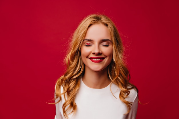 Chica romántica de pelo largo posando con sonrisa tímida. Foto interior de atractiva mujer europea aislada en la pared roja.