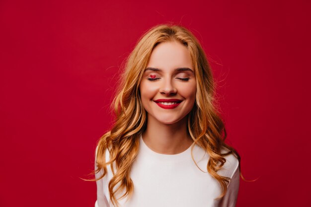 Chica romántica de pelo largo posando con sonrisa tímida. Foto interior de atractiva mujer europea aislada en la pared roja.