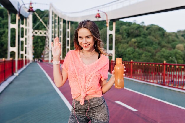 Chica romántica en rosa t-short divirtiéndose en el estadio. Mujer joven adorable con una botella de agua riéndose de la pista de ceniza.