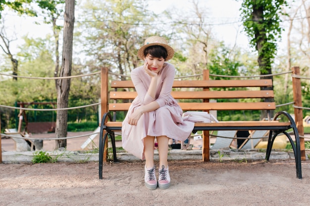 Chica romántica con sombrero de paja de moda sentado en un banco del parque apuntando la cara con la mano y pensando en algo bueno