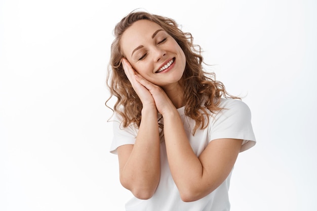 Una chica romántica sonriente soñando despierta recuerda un momento hermoso y feliz cierra los ojos e imagina algo apoyado en las manos durmiendo de ensueño de pie sobre un fondo blanco