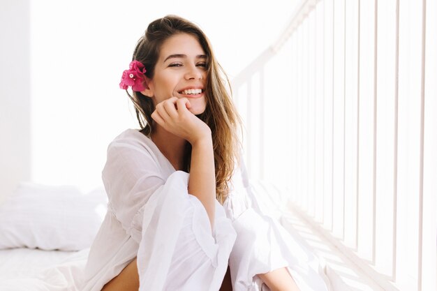 Chica romántica con sonrisa astuta en blusa vintage sentada en la cama y tocando su barbilla con la mano. Retrato de mujer joven linda soñadora con flor en peinado descansando en el dormitorio en la mañana