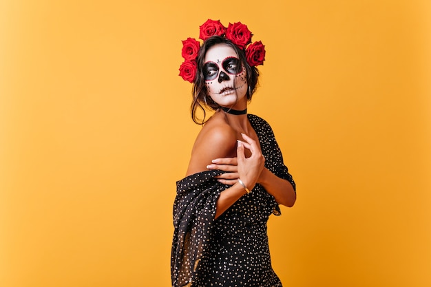 La chica romántica sostiene suavemente el vestido que cae. Señora con maquillaje en forma de calavera para carnaval misteriosamente mira hacia otro lado.