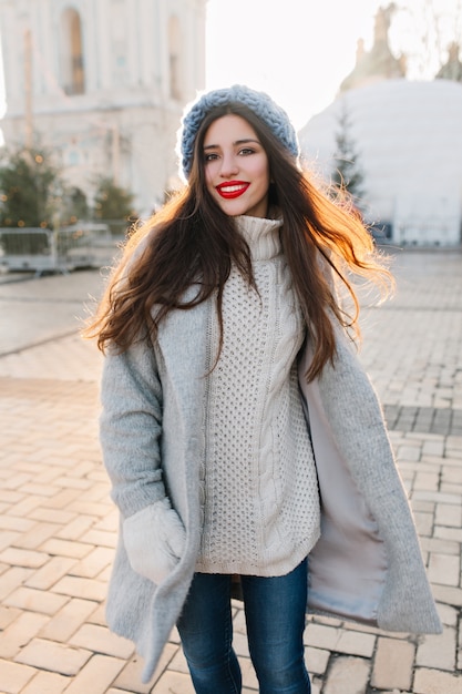 Foto gratuita chica romántica en suéter de punto largo posando con una sonrisa durante la caminata matutina en diciembre. mujer morena guapa en abrigo gris y jeans escalofriantes en la ciudad en invierno.