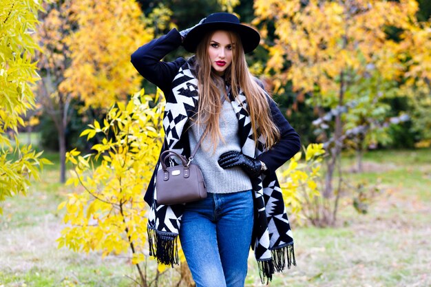 Chica romántica en vasos con sombrero y posando con expresión de cara de besos de pie en medio del parque. Retrato al aire libre de linda mujer joven con bufanda de moda divirtiéndose durante la caminata en el bosque.