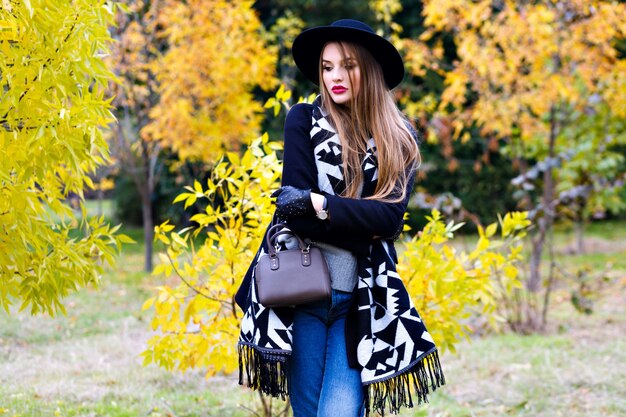 Chica romántica en vasos con sombrero y posando con expresión de cara de besos de pie en medio del parque. Retrato al aire libre de linda mujer joven con bufanda de moda divirtiéndose durante la caminata en el bosque.