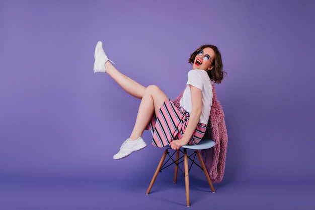 Chica romántica en zapatos blancos sentada en una silla y riendo. Filmación en interiores de mujer rizada sin preocupaciones posando en la pared púrpura con una sonrisa sincera.