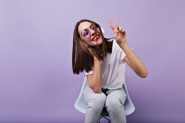 Chica en ropa blanca de moda posando con el signo de la paz mientras escucha música. Impresionante mujer en auriculares y gafas de sol sonriendo.