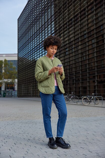 Chica con ropa elegante sostiene un moderno dispositivo de teléfono inteligente para comunicarse intenta encontrar rutas, descargas, aplicaciones de navegación, se encuentra en un entorno urbano