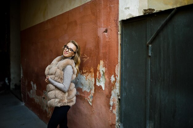 Chica rubia con abrigo de piel y gafas posadas contra la vieja pared naranja