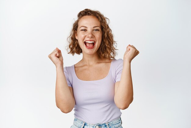 Chica rubia alegre celebrando cantando y gritando con alegría ganando algo de pie contra el fondo blanco.