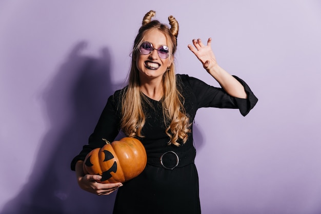 Chica rubia alegre en traje de bruja disfrutando del carnaval. Filmación en interiores de dama despreocupada sonriente con pie de calabaza de halloween en la pared púrpura.