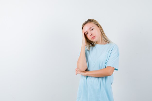 Chica rubia apoyada en la palma de la mano, sosteniendo la mano debajo del codo mientras posa con una camiseta azul y se ve bonita. vista frontal.