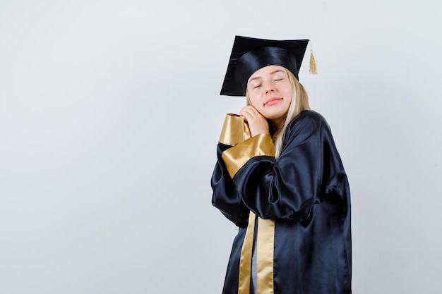 Chica rubia apoyando la mejilla en las manos, cerrando los ojos en toga y gorra de graduación y mirando soñoliento