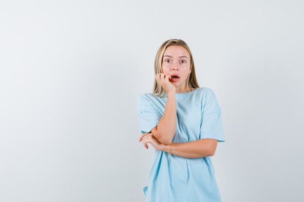 Chica rubia apoyando la mejilla en la palma en camiseta azul y mirando sorprendido, vista frontal.