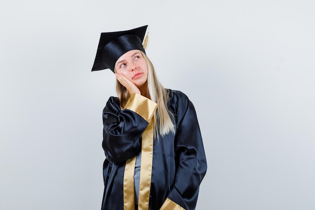 Chica rubia apoyándose en la palma de la mano en toga y gorra de graduación y mirando pensativo.