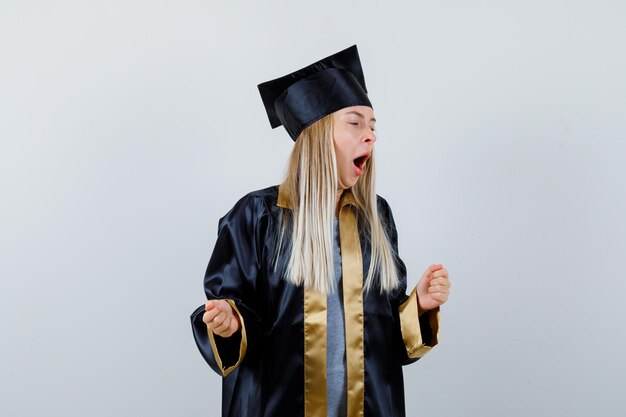 Chica rubia apretando los puños, abriendo la boca en toga y gorra de graduación y con aspecto soñoliento