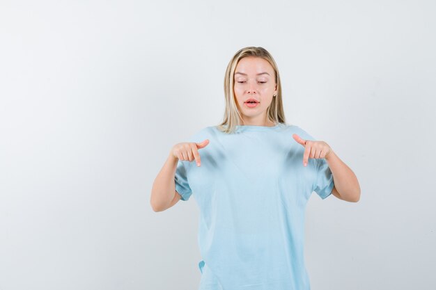 Chica rubia apuntando hacia abajo con los dedos índices en camiseta azul y mirando enfocado. vista frontal.