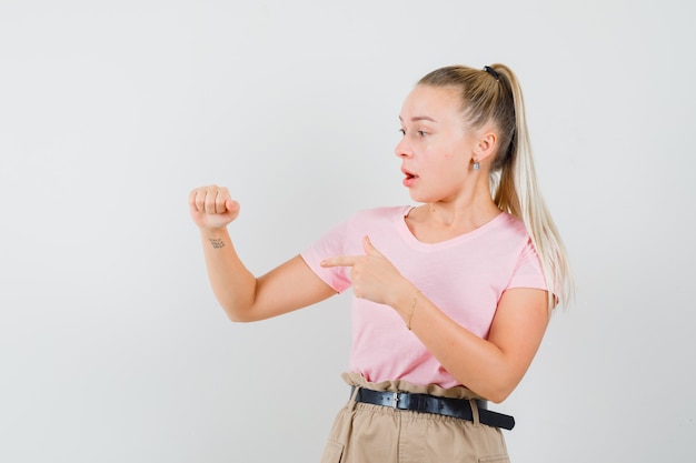 Chica rubia apuntando a algo fingió estar retenida en camiseta, pantalones y mirando sorprendida. vista frontal.