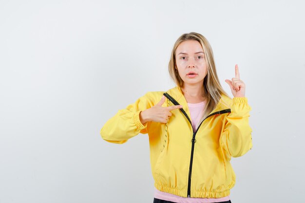 Chica rubia apuntando hacia arriba y hacia la derecha con los dedos índices en camiseta rosa y chaqueta amarilla y mirando seria.