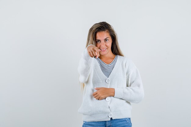 Chica rubia apuntando a la cámara con camiseta a rayas, cárdigan blanco y pantalones de mezclilla y mirando feliz. vista frontal.