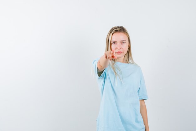 Chica rubia apuntando a la cámara con el dedo índice en camiseta azul y mirando serio, vista frontal.