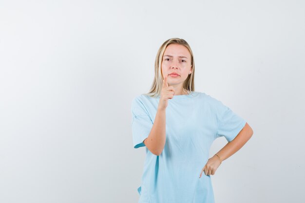 Chica rubia apuntando a la cámara con el dedo índice, sosteniendo la mano en la cintura en una camiseta azul y mirando seria, vista frontal.