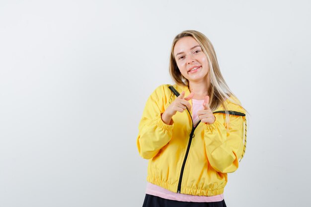 Chica rubia apuntando a la cámara con los dedos índices en camiseta rosa y chaqueta amarilla y mirando seria