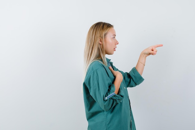 Chica rubia apuntando a la derecha mientras sostiene la mano en el pecho con una blusa verde y mira concentrada.