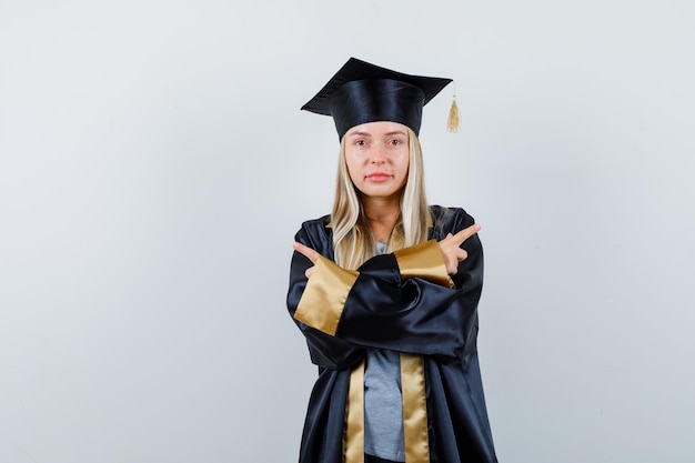 Chica rubia apuntando en direcciones opuestas en toga y gorra de graduación y mirando seria.