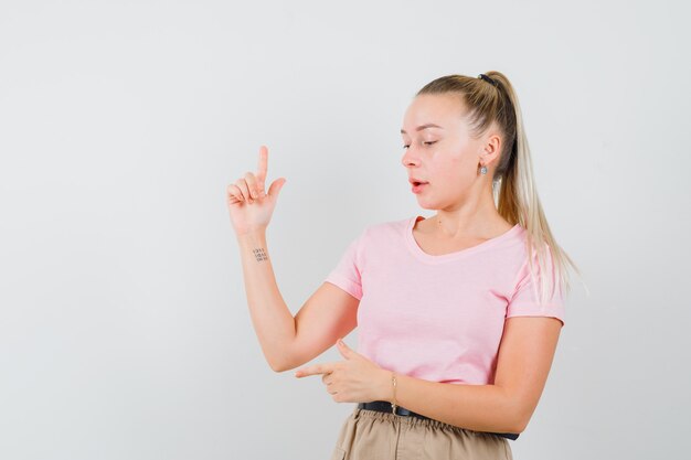 Chica rubia apuntando hacia un lado y hacia arriba en camiseta, pantalones y mirando enfocado, vista frontal.