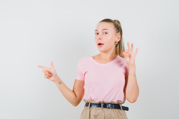 Chica rubia apuntando a un lado, mostrando un gesto aceptable en camiseta, pantalones y luciendo confiada. vista frontal.