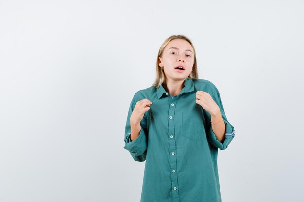 Chica rubia apuntando a sí misma en blusa verde y mirando sorprendida