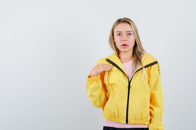 Chica rubia apuntando a sí misma en camiseta rosa y chaqueta amarilla y luciendo seductora.