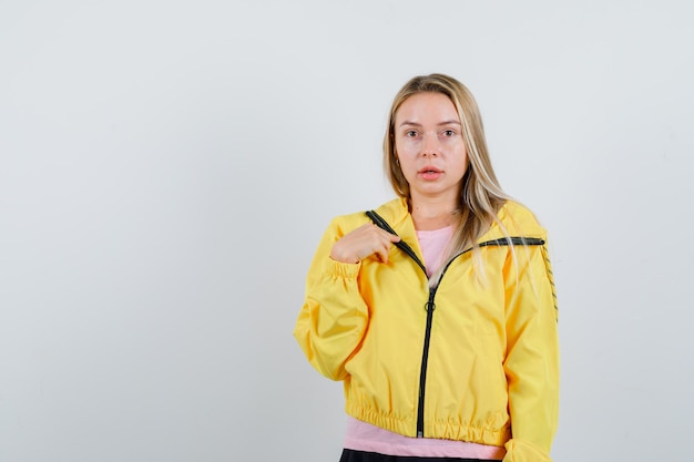 Chica rubia apuntando a sí misma en chaqueta amarilla y mirando perpleja