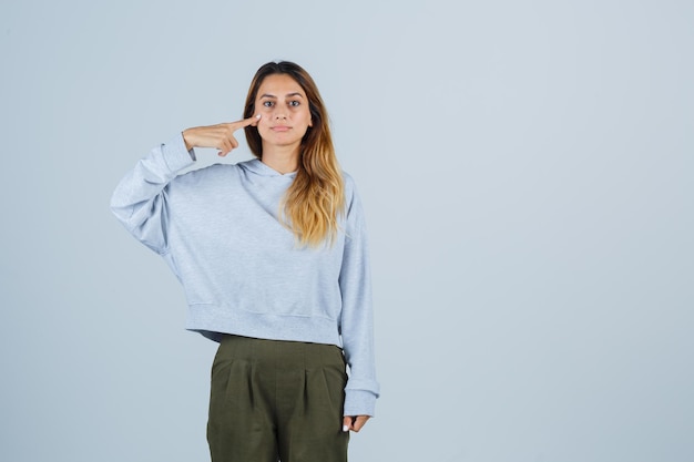 Chica rubia apuntando a sí misma con el dedo índice en sudadera azul verde oliva y pantalones y mirando seria. vista frontal.