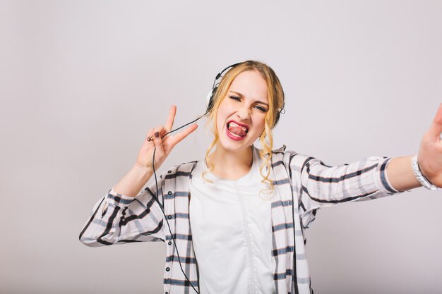 Chica rubia en auriculares con la lengua fuera disfruta de la música y bromea bailando aislado. Mujer joven atractiva con el pelo rizado escuchando su canción favorita y divirtiéndose