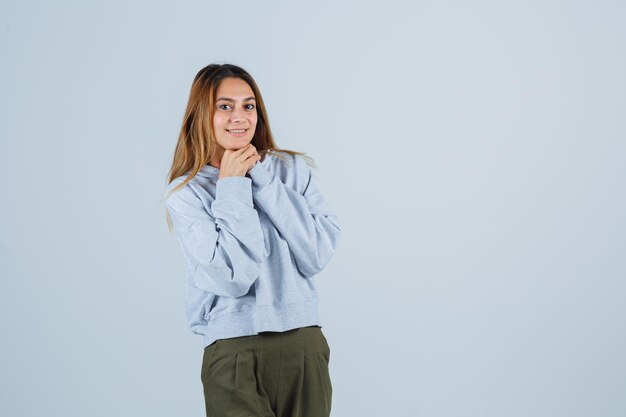 Chica rubia con la barbilla apoyada en las manos, uniendo las manos en pantalones y sudadera azul verde oliva y mirando feliz. vista frontal.