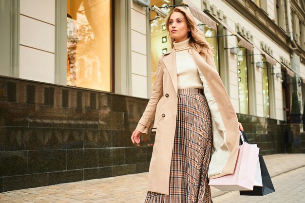 Chica rubia bastante elegante con abrigo beige con bolsas de compras caminando por la calle de la ciudad