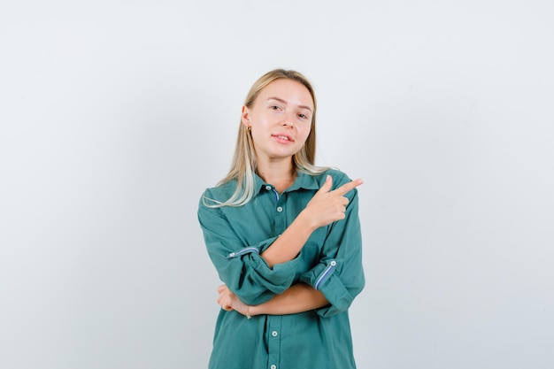 Chica rubia en blusa verde apuntando a la derecha con el dedo índice y luciendo bonita